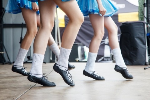 Legs and feet of 3 Irish step dancers wearing blue skirts, white socks, and black soft soled dancing shoes.